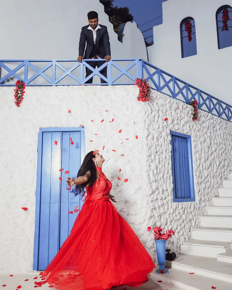 Woman in a long red gown on white steps, rose petals falling.