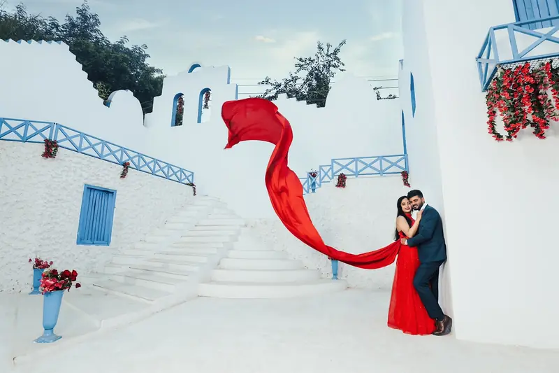 Couple on a white staircase, the bride's red dupatta lifted by the wind.